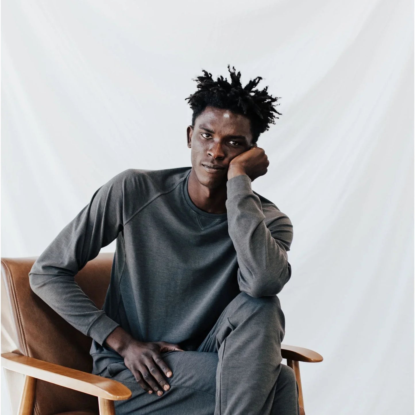 Man in gray casual outfit sitting on brown chair against white backdrop, thoughtful pose. Come see us at Awaken, Franklin, Tennessee