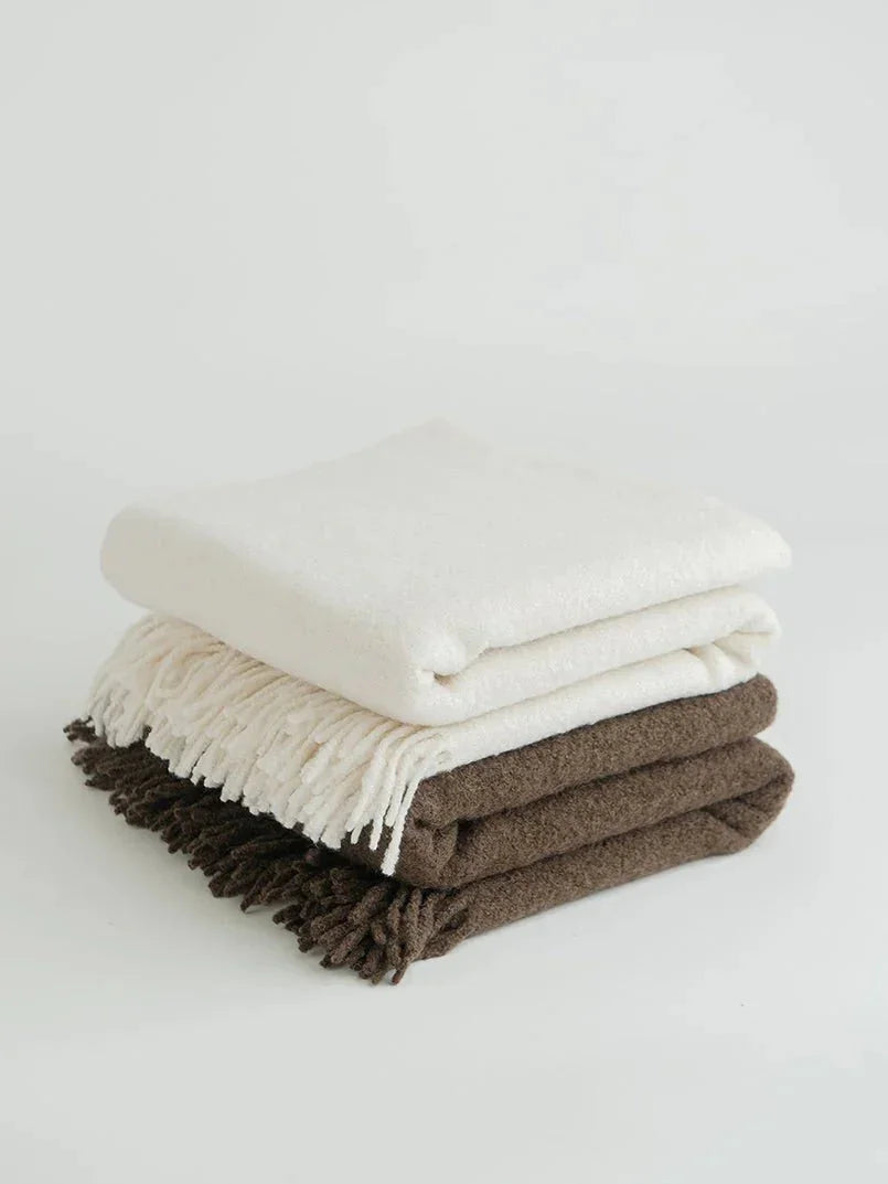 Stack of soft white and brown blankets with fringes on a white background. Come see us at Awaken, Franklin, Tennessee