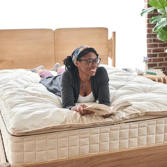 Smiling woman reading on a plush beige mattress in a bright bedroom with wooden bed frame. Come see us at Awaken, Franklin, Tennessee