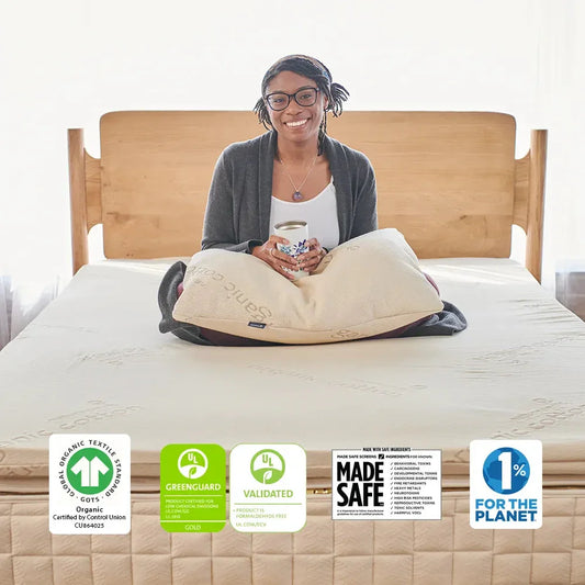 Smiling woman sitting cross-legged on an organic mattress holding a cup with a pillow, wooden headboard in bright room. Come see us at Awaken, Franklin, Tennessee