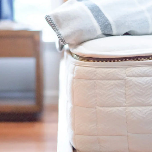 Close-up of white quilted mattress corner with zipper and gray-striped blanket, wooden furniture blurred. Come see us at Awaken, Franklin, Tennessee