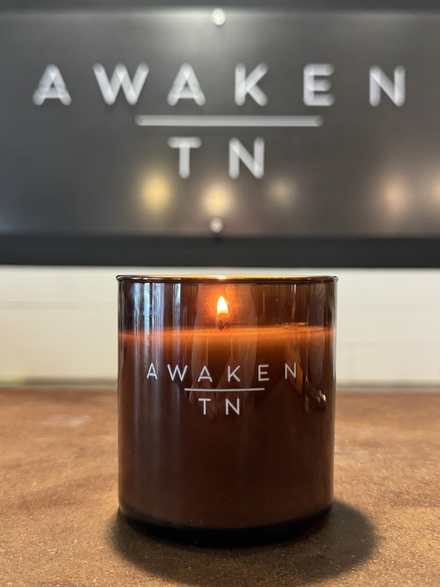 Lit Awaken TN candle on wooden surface, branded sign in background. Come see us at Awaken, Franklin, Tennessee.