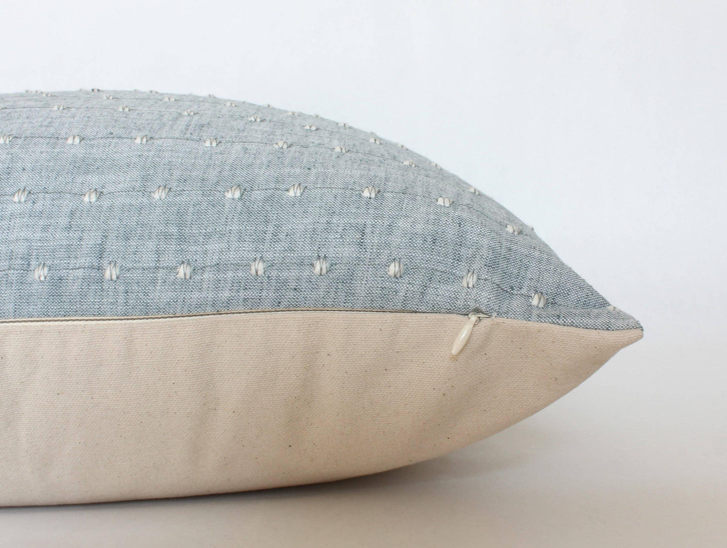Close-up of blue textured pillow with white stitched dots and beige fabric bottom with zipper, on white background. Come see us at Awaken, Franklin, Tennessee