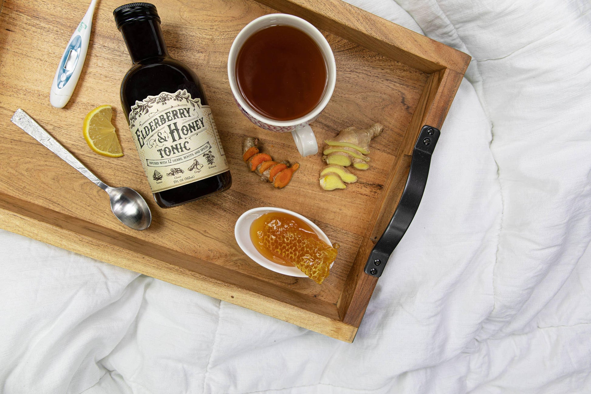 Wood tray with elderberry & honey tonic, tea cup, honeycomb, ginger, turmeric, lemon slice on white blanket. Come see us at Awaken, Franklin, Tennessee