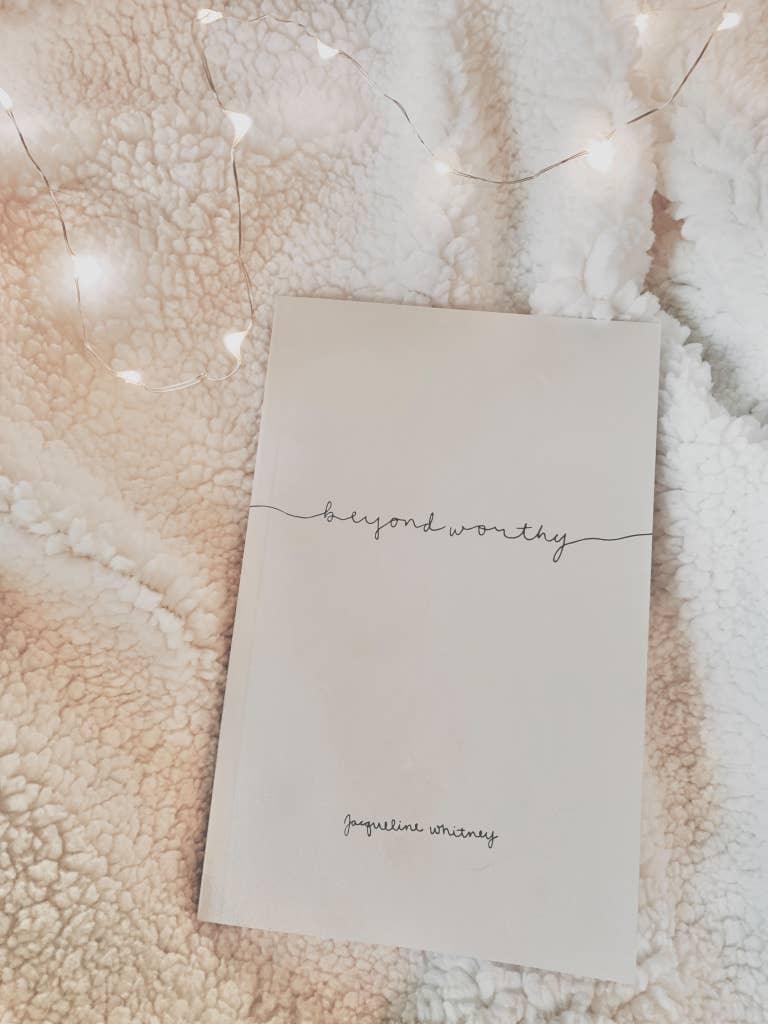 Book titled Beyond Worthy by Jaqueline Whitney on textured white blanket with string lights, cozy setting. Come see us at Awaken, Franklin, Tennessee