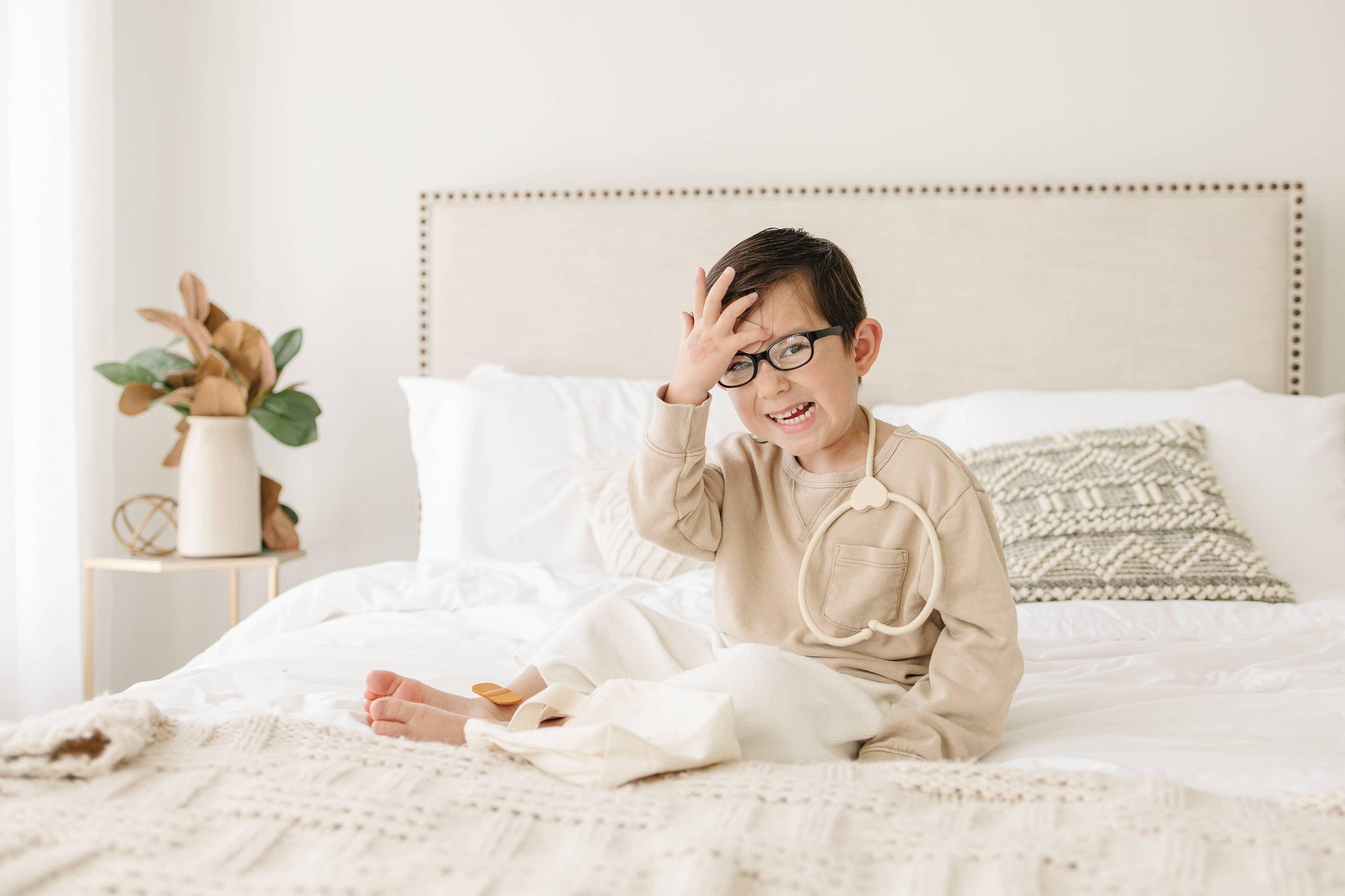 Smiling young boy with glasses sitting on white bed in beige room with decorative pillow and plant. Come see us at Awaken, Franklin, Tennessee