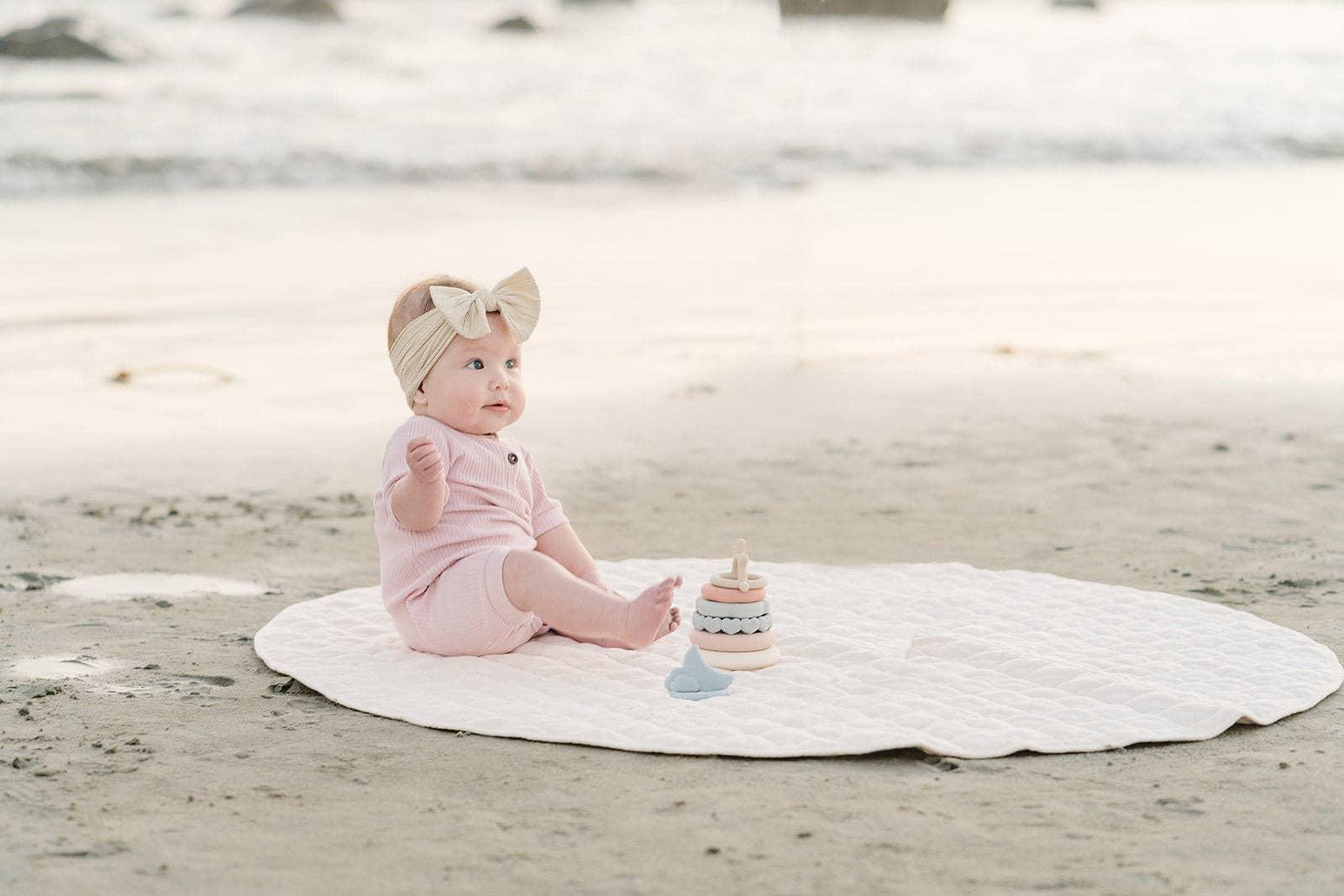 Baby in pink outfit with beige headband sitting on white mat at beach with toy. Come see us at Awaken, Franklin, Tennessee