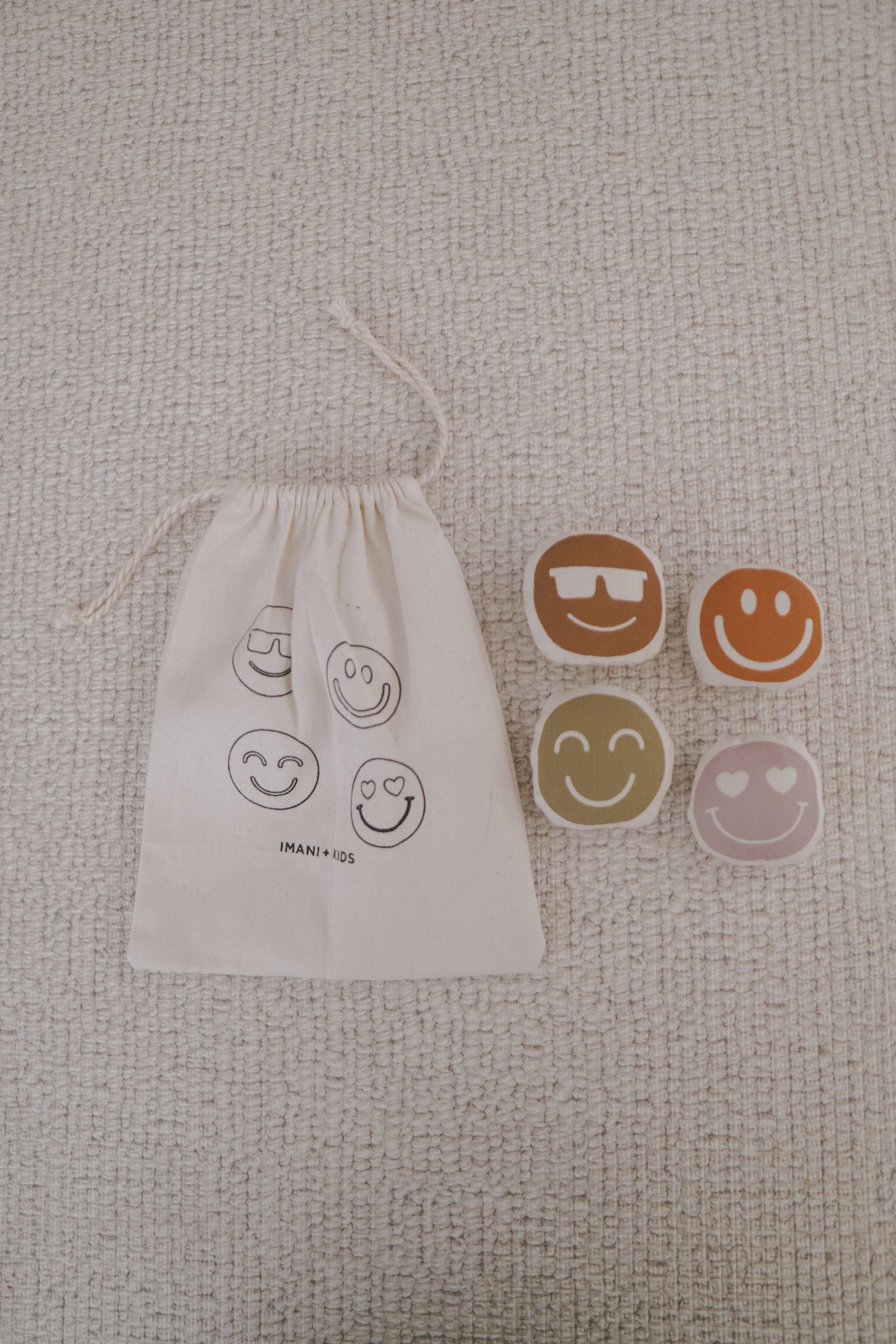 Imani + Kids drawstring bag with smiley face plush toys on beige textured surface, Come see us at Awaken, Franklin, Tennessee