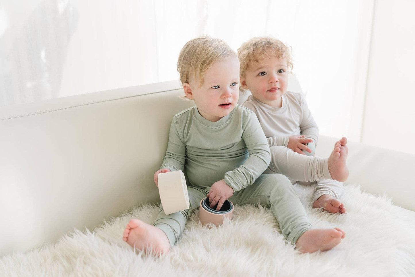 Two toddlers in pastel loungewear sitting on a white fluffy rug indoors, light-filled room. Come see us at Awaken, Franklin, Tennessee