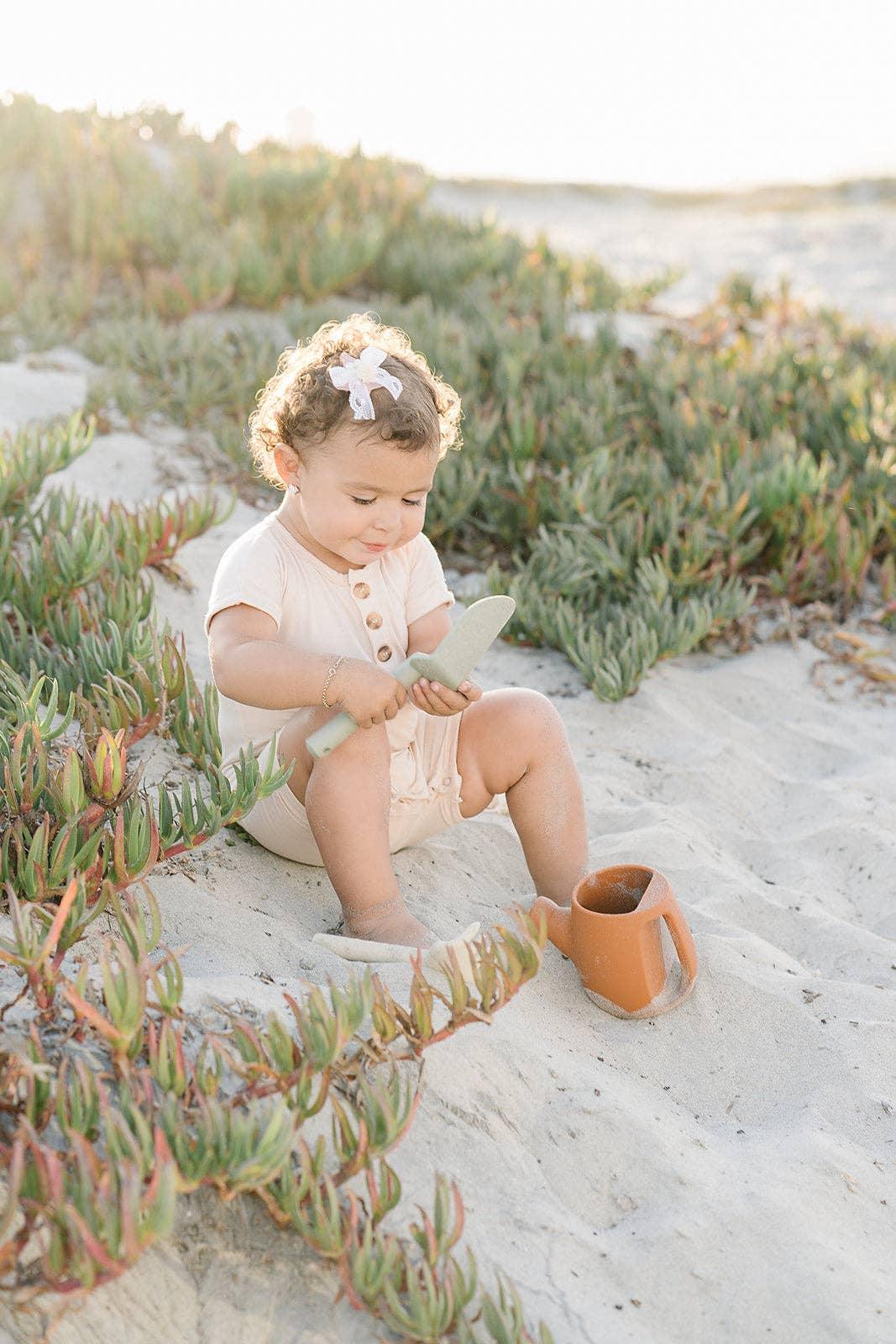 Toddler with curly hair and bow sitting on sandy beach near greenery, playing with green toy shovel and orange bucket. Come see us at Awaken, Franklin, Tennessee