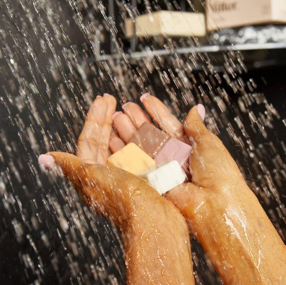 Hands holding colorful soap bars under running water with soap shelves in background. Come see us at Awaken, Franklin, Tennessee