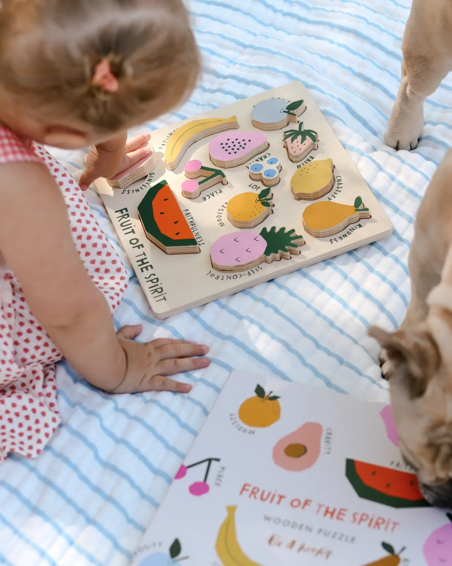 Child playing with colorful wooden Fruit of the Spirit puzzle on a striped blanket with a dog nearby. Come see us at Awaken, Franklin, Tennessee