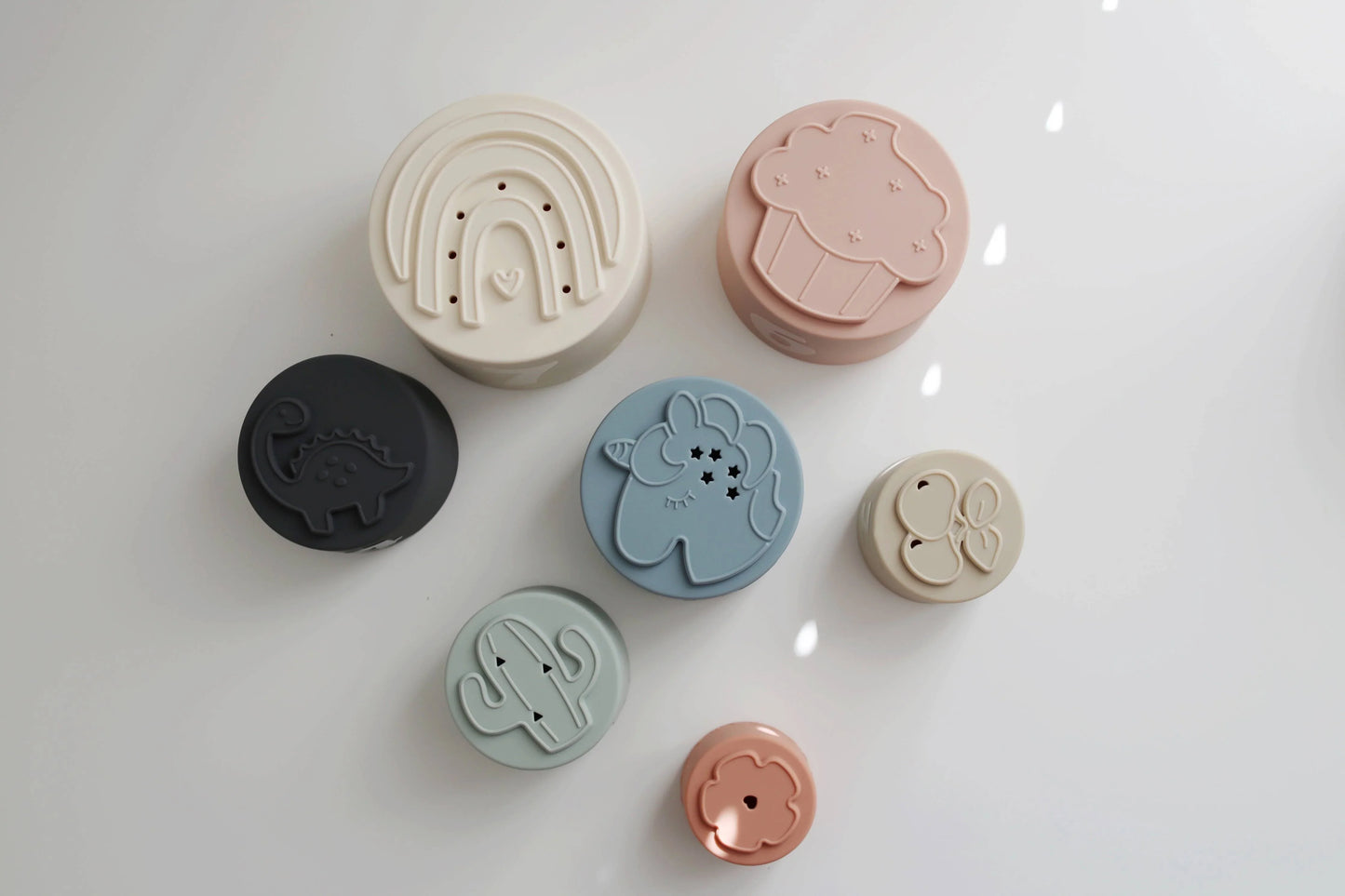 Six pastel silicone objects with embossed designs including rainbow, cupcake, dinosaur, unicorn, cactus, and cherries on white surface. Come see us at Awaken, Franklin, Tennessee