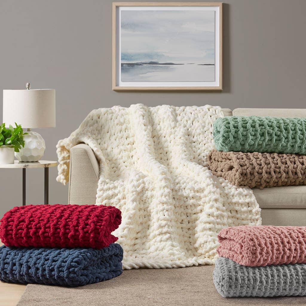 Chunky knit blankets in cream, green, brown, red, blue, pink, and gray on sofa and floor with lamp and plant; Come see us at Awaken, Franklin, Tennessee