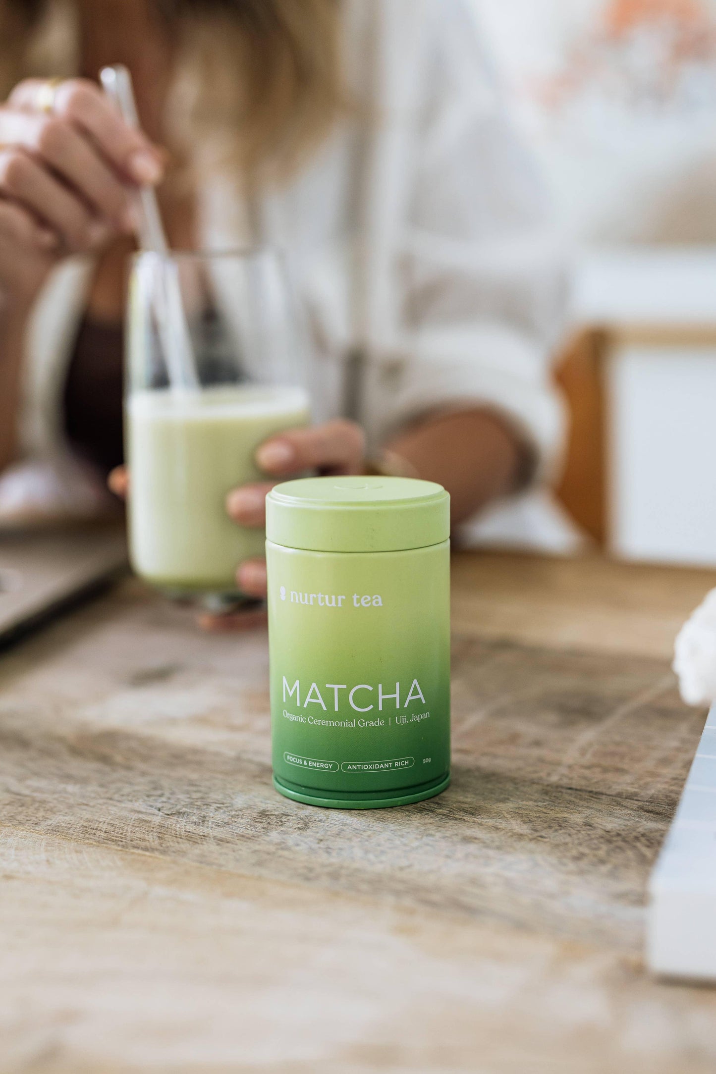 Green Nurtur Tea Matcha container on a wooden table with a woman stirring a glass of matcha latte. Come see us at Awaken, Franklin, Tennessee
