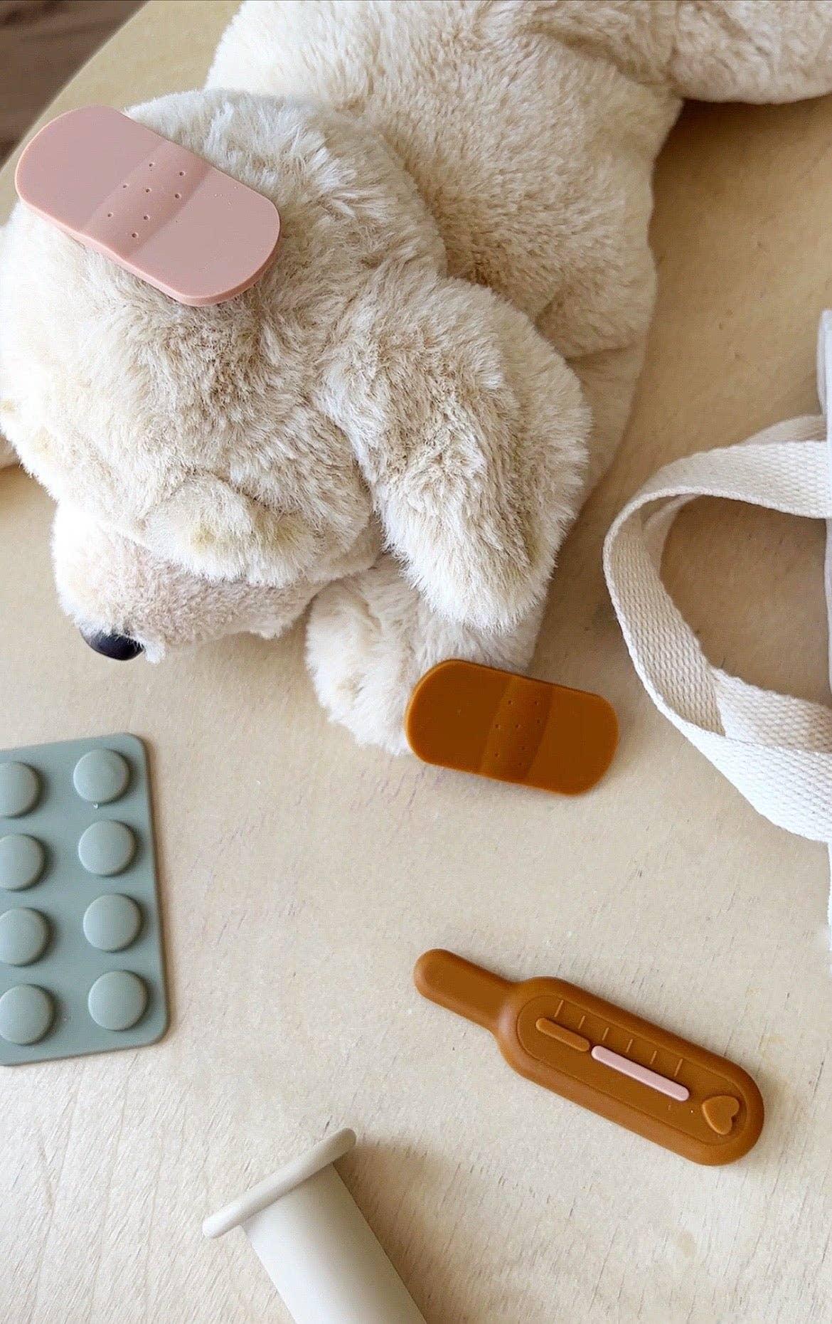 Plush teddy bear with toy bandages and thermometer on beige surface; children's medical playset. Come see us at Awaken, Franklin, Tennessee.