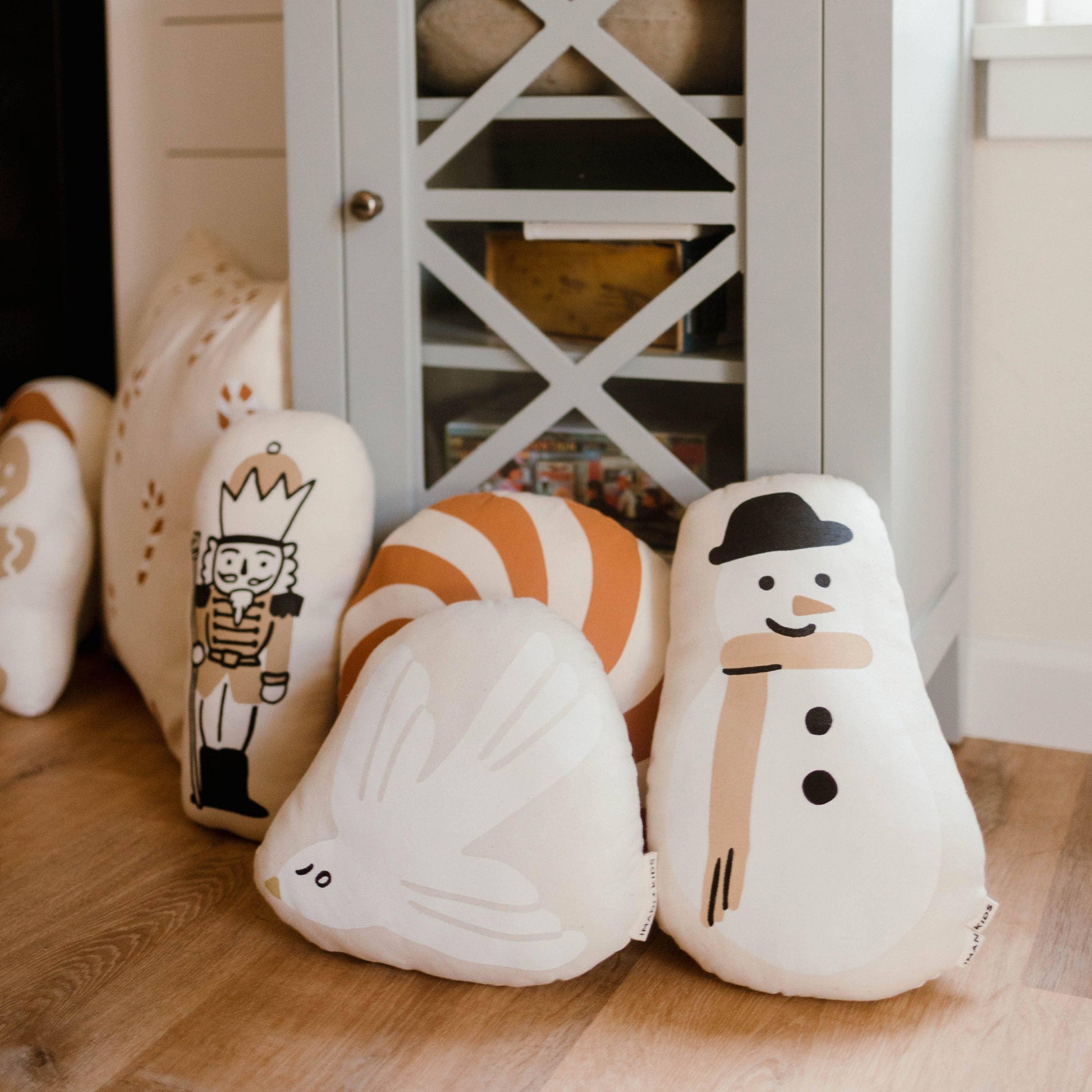 Festive holiday pillows with nutcracker, snowman, bird, and candy designs on wood floor, cabinet in background. Come see us at Awaken, Franklin, Tennessee.