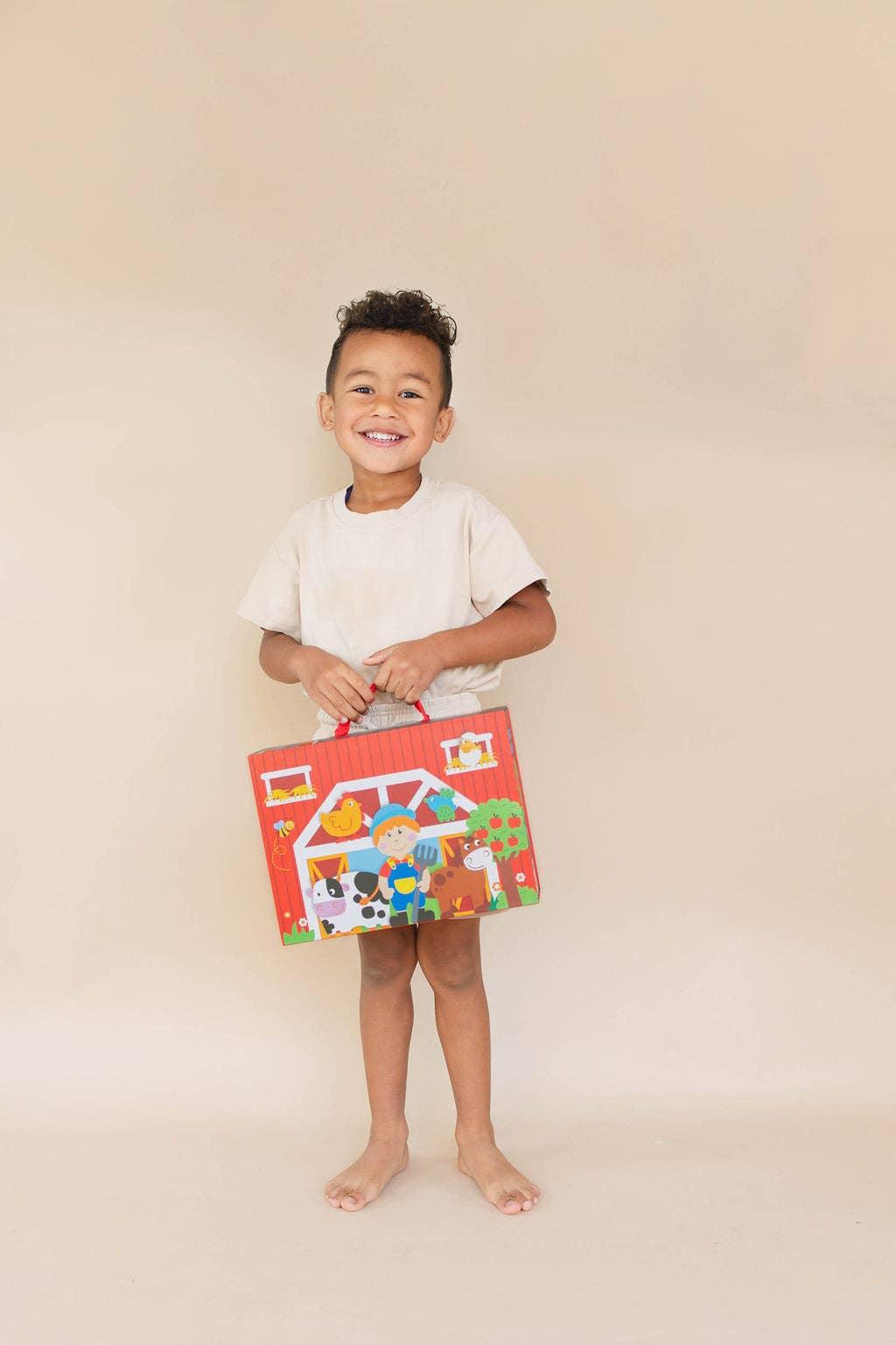Smiling boy in beige clothes holding colorful barnyard toy box with farm animals, plain background. Come see us at Awaken, Franklin, Tennessee