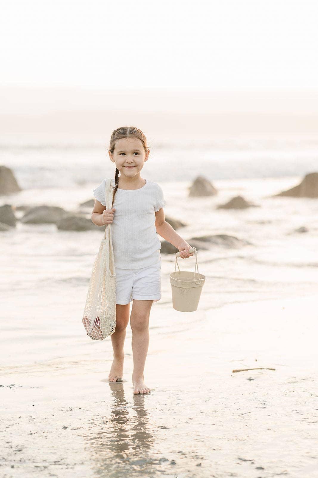 Young girl in white outfit walking barefoot on beach holding bucket and tote bag. Come see us at Awaken, Franklin, Tennessee