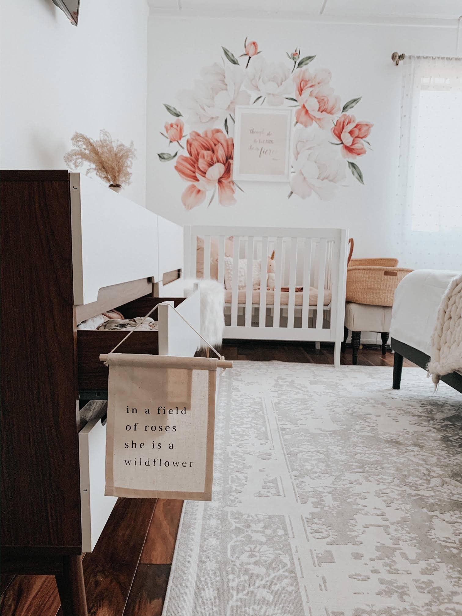 Neutral nursery with crib, floral wall art, sign reading 'in a field of roses she is a wildflower,' cozy decor—Come see us at Awaken, Franklin, Tennessee.