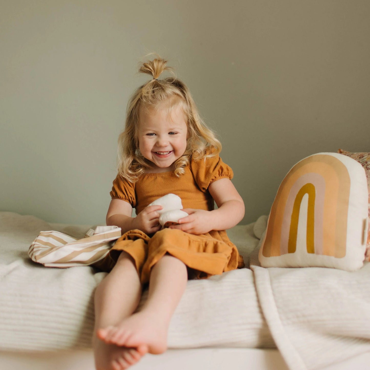 Smiling blonde toddler in mustard dress with rainbow pillow on cozy bed. Come see us at Awaken, Franklin, Tennessee