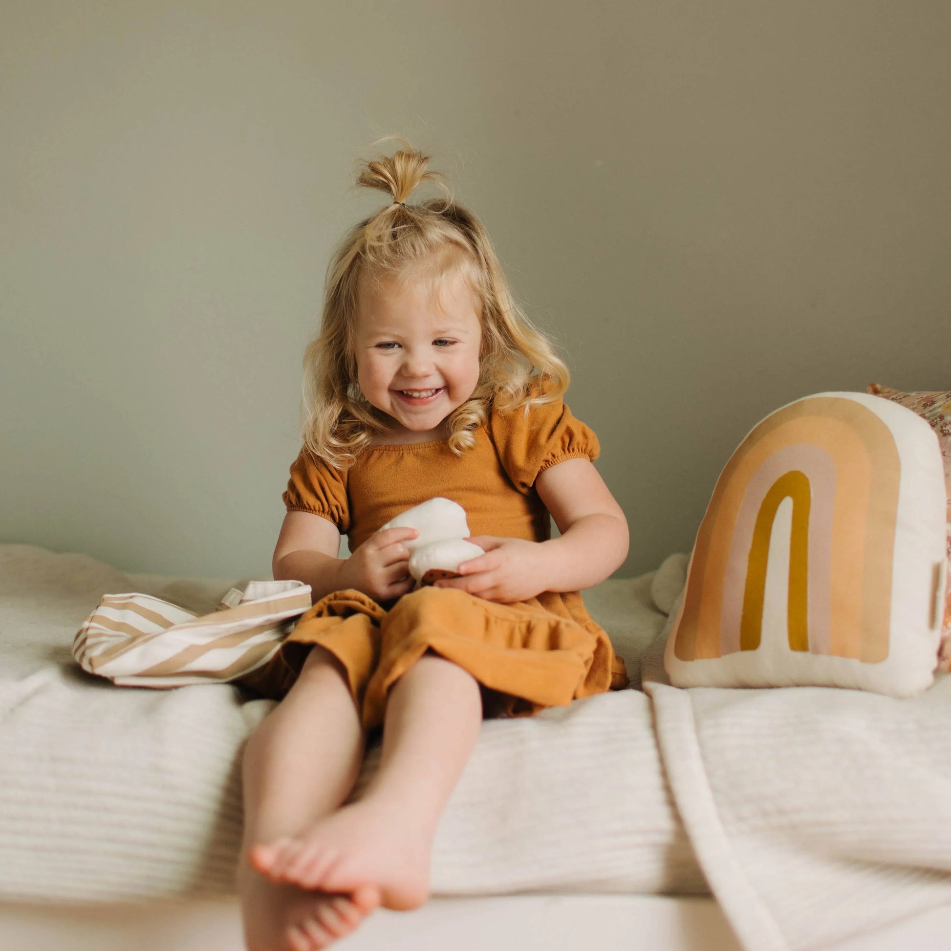 Smiling blonde toddler in mustard dress with rainbow pillow on cozy bed. Come see us at Awaken, Franklin, Tennessee