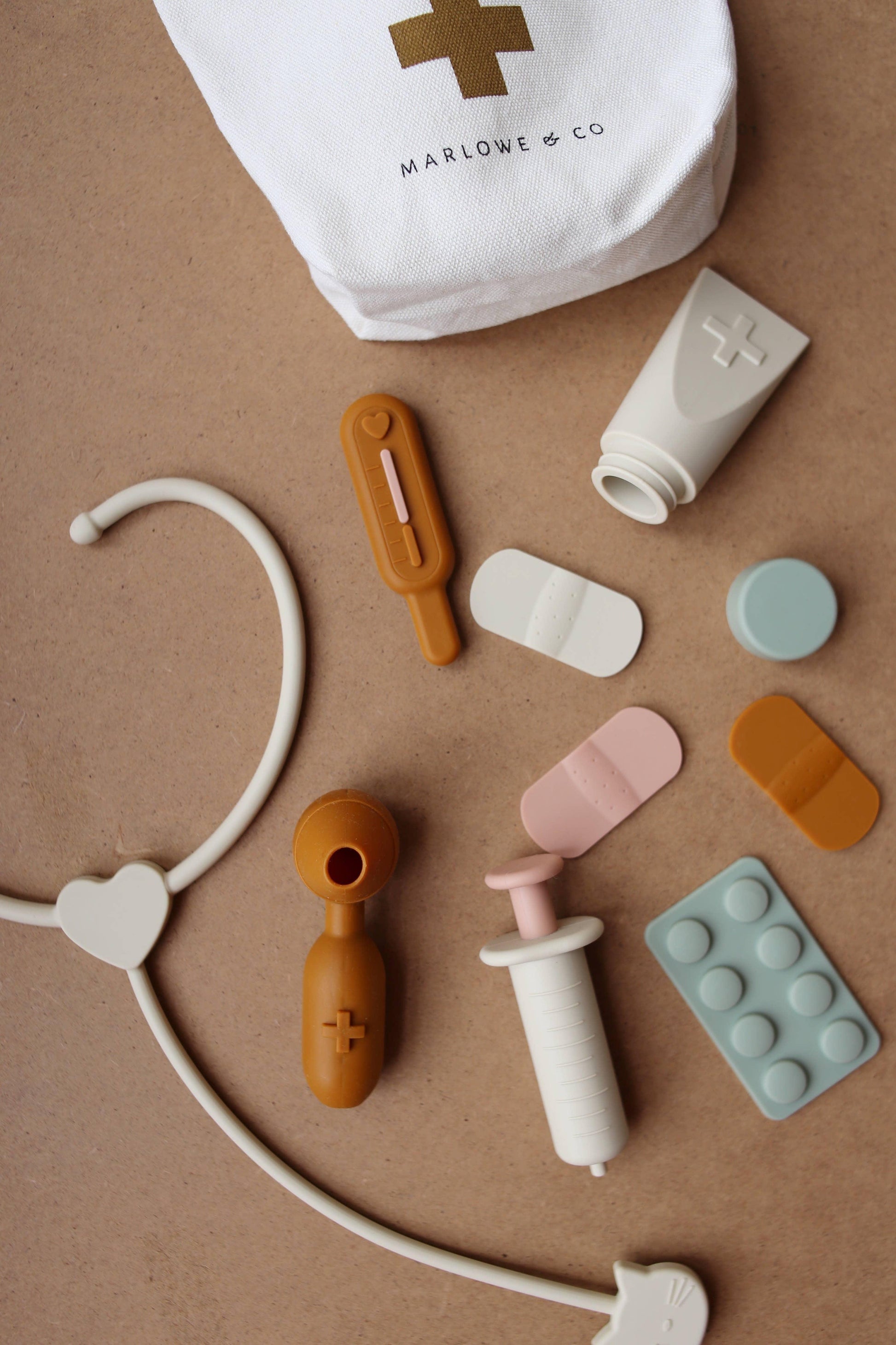 Toy medical kit with stethoscope, syringe, thermometer, bandages, and Marlowe & Co bag on brown surface. Come see us at Awaken, Franklin, Tennessee