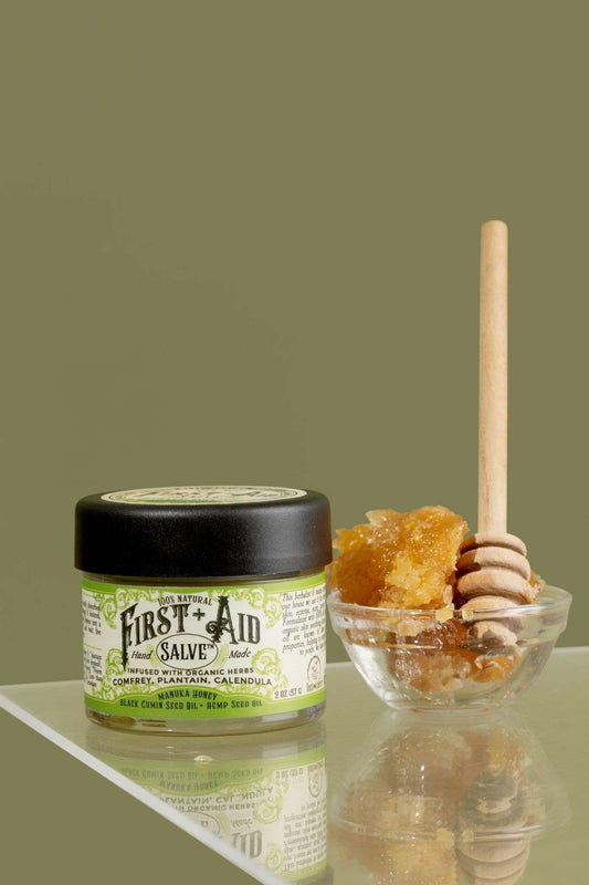 Jar of First Aid Salve with organic herbs next to honeycomb and wooden dipper on glass surface. Come see us at Awaken, Franklin, Tennessee