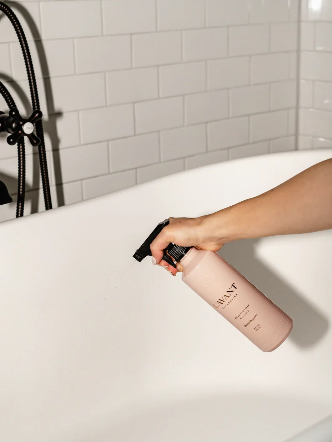 Hand spraying pink cleaner bottle on white bathtub, tile wall background. Come see us at Awaken, Franklin, Tennessee