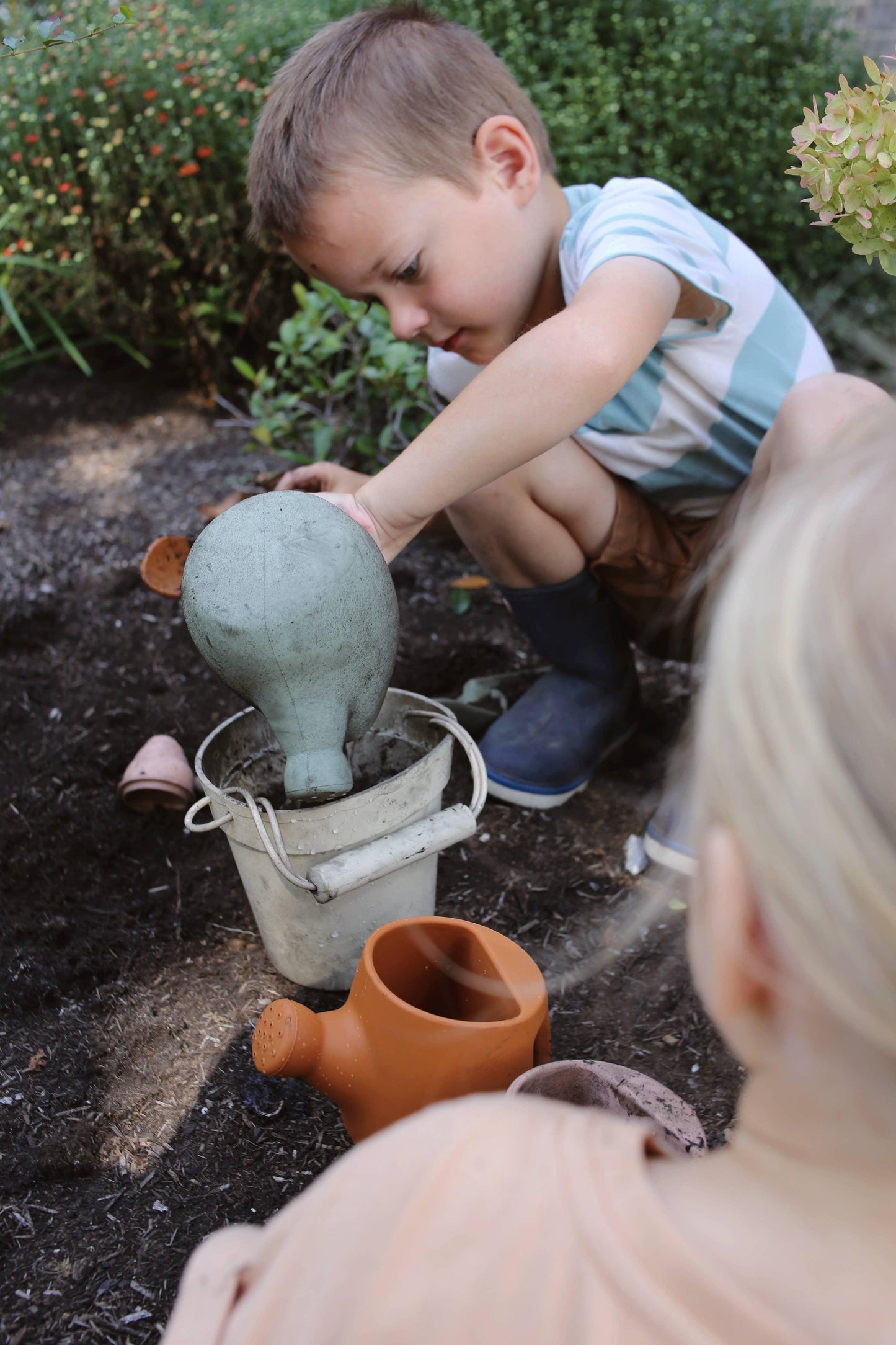 Young boy pouring water from green watering can into bucket in garden with plants. Come see us at Awaken, Franklin, Tennessee
