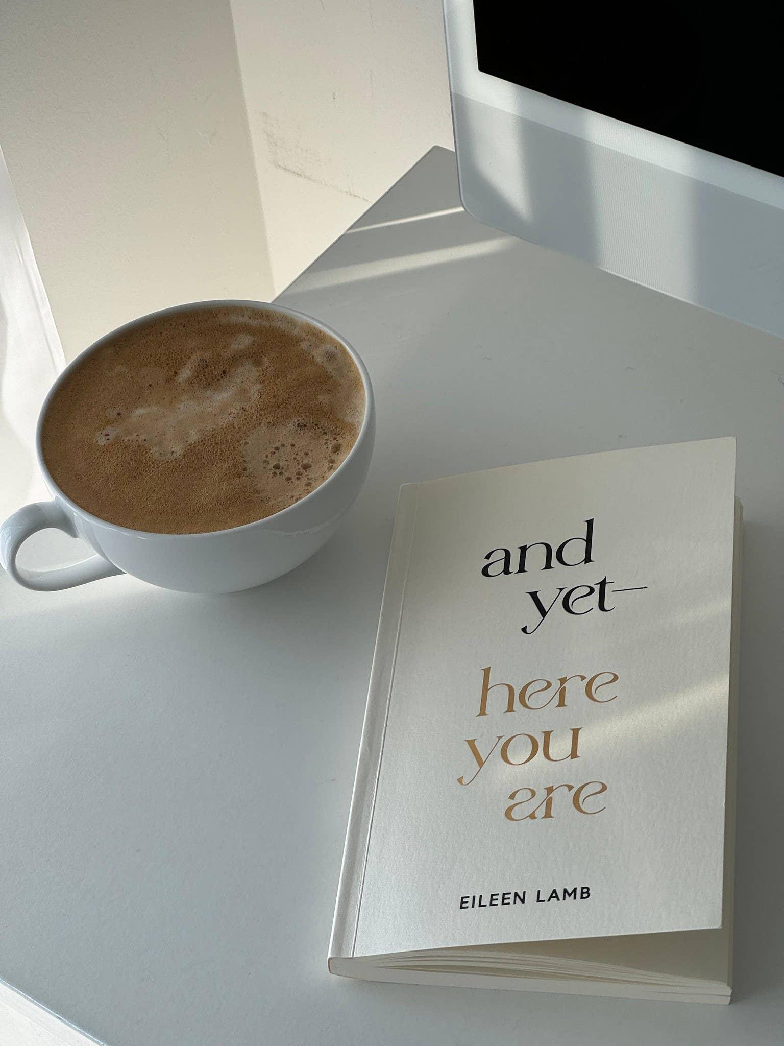 Cup of coffee beside the book 'and yet here you are' by Eileen Lamb on a white desk. Come see us at Awaken, Franklin, Tennessee