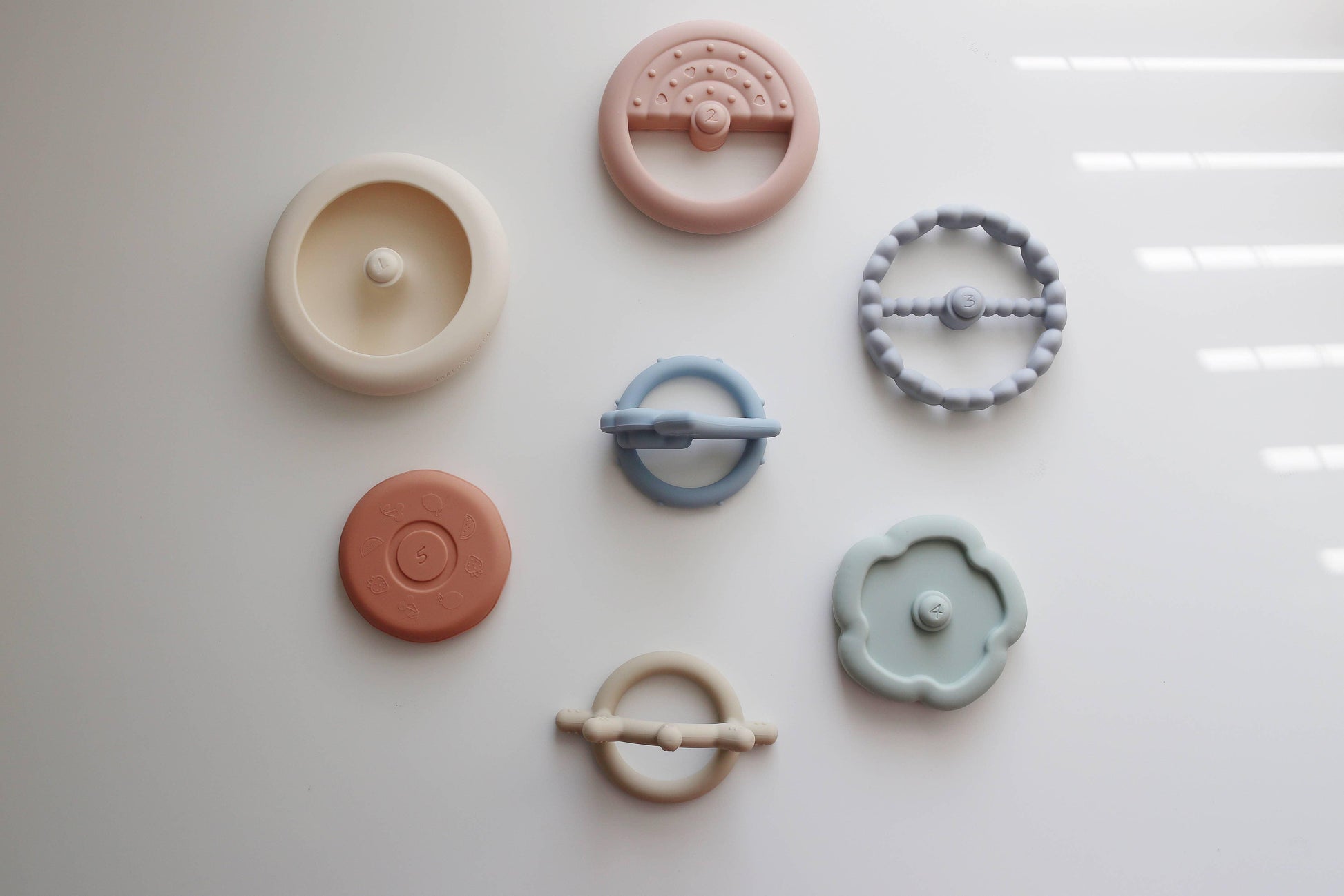 Set of seven pastel-colored silicone baby teethers laid out on white surface. Come see us at Awaken, Franklin, Tennessee