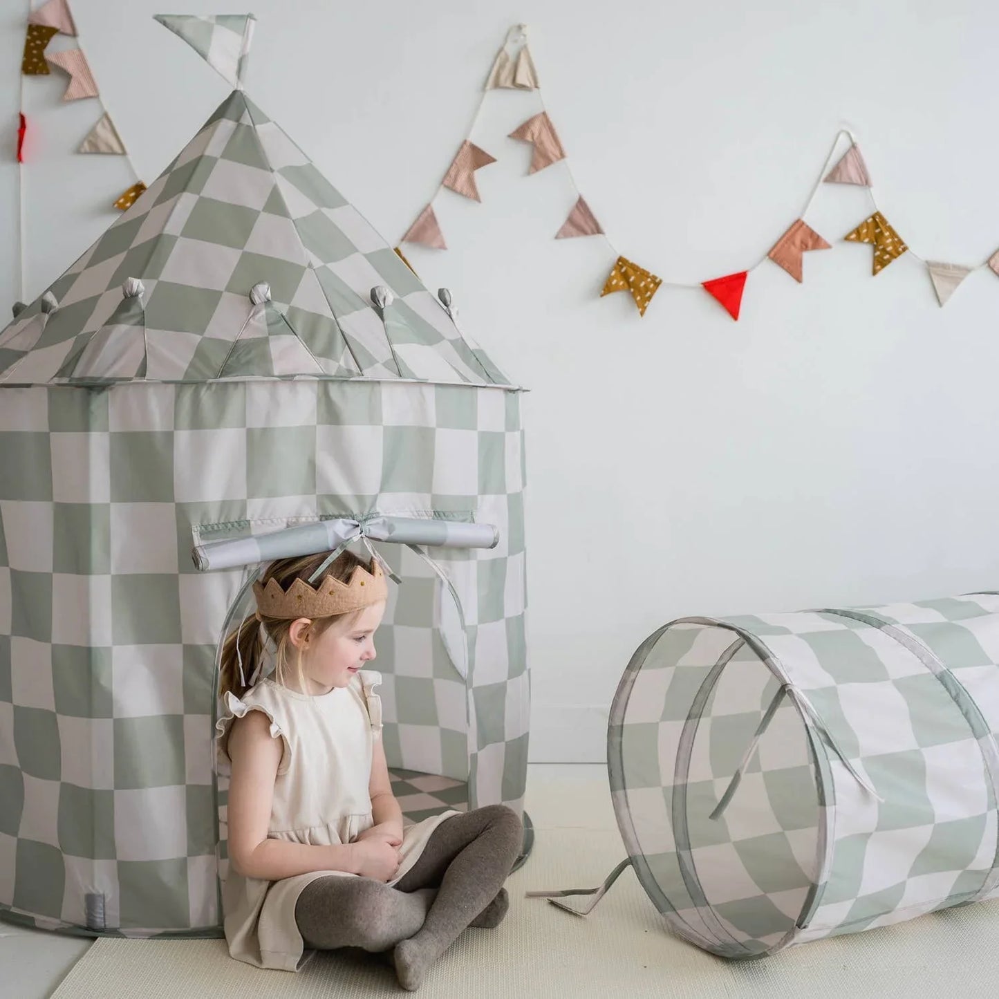 Young girl wearing a crown sitting beside a gray and white checkered play tent and tunnel with colorful bunting on wall. Come see us at Awaken, Franklin, Tennessee