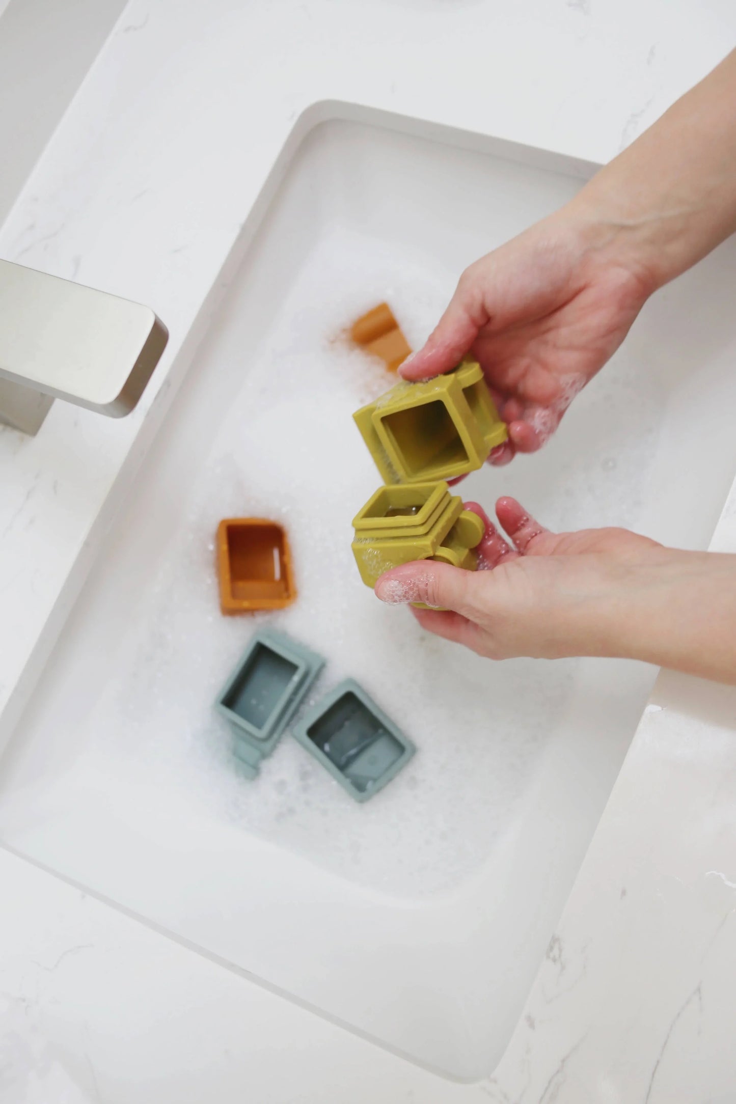 Hands washing colorful silicone baby toys in bubbly water sink, white marble countertop. Come see us at Awaken, Franklin, Tennessee