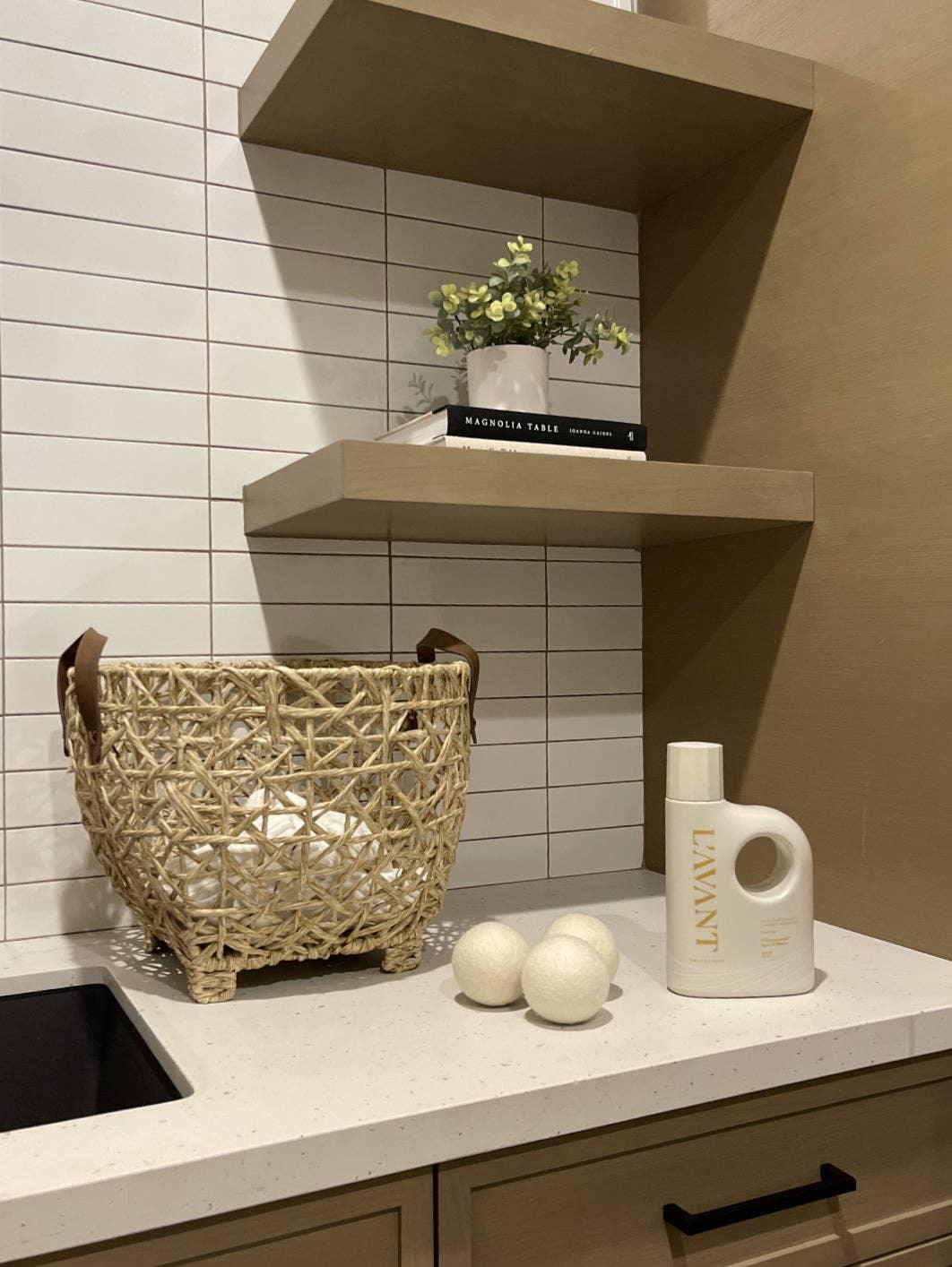 Laundry room with woven basket, laundry detergent, dryer balls, plant decor, and books on shelves. Come see us at Awaken, Franklin, Tennessee.
