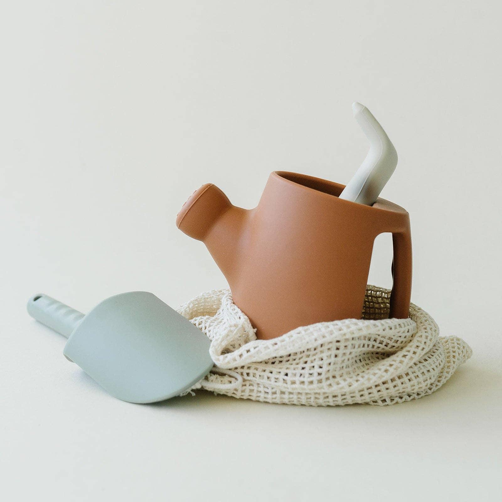 Terracotta watering can and gray gardening spatula on white mesh bag, minimal gardening tools. Come see us at Awaken, Franklin, Tennessee