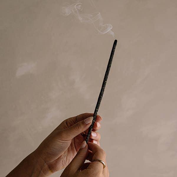 Hands holding a lit incense stick with smoke against a beige background. Come see us at Awaken, Franklin, Tennessee