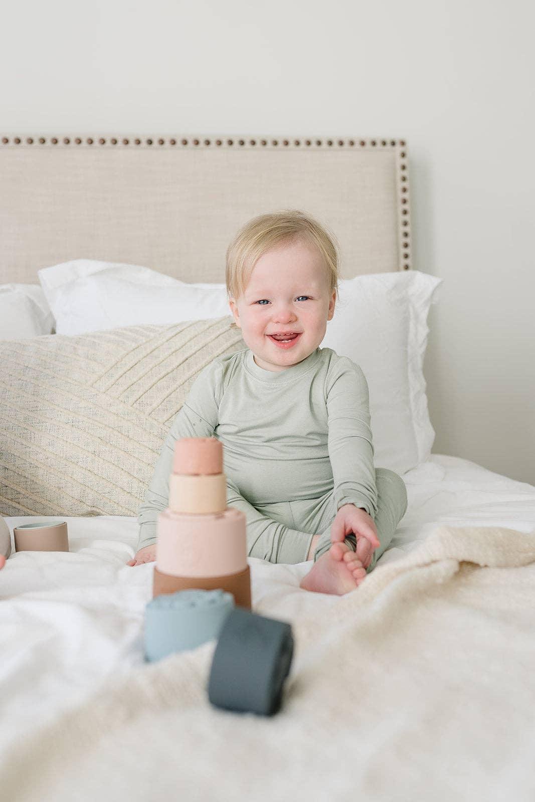 Smiling toddler in light green pajamas sitting on bed playing with pastel stacking cups. Come see us at Awaken, Franklin, Tennessee