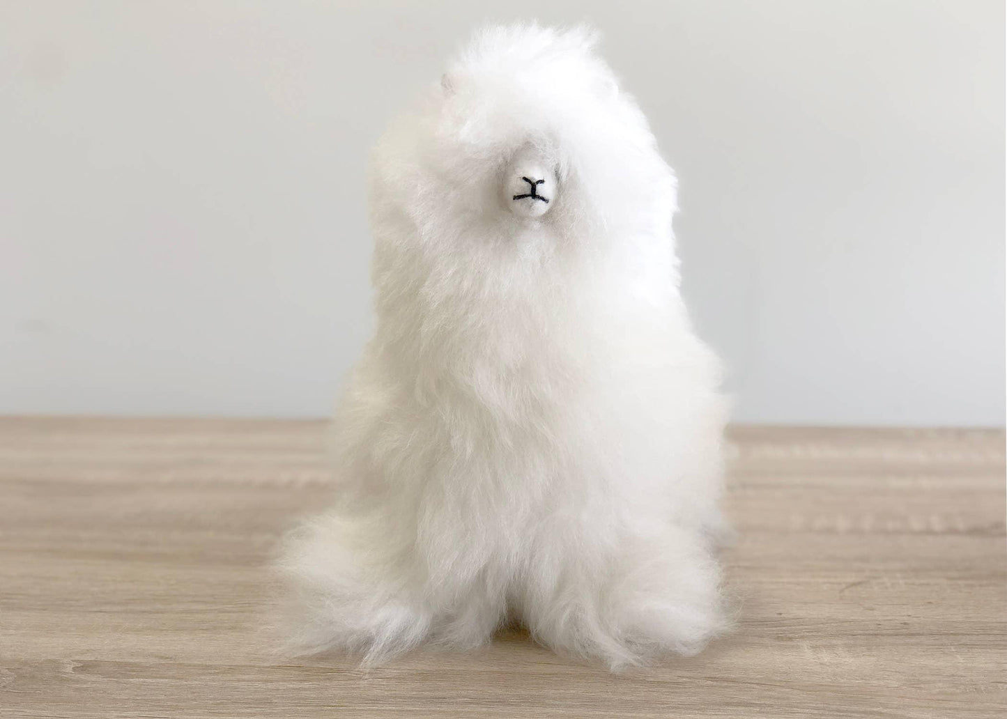 White fluffy alpaca plush toy on wooden table with light gray background. Come see us at Awaken, Franklin, Tennessee