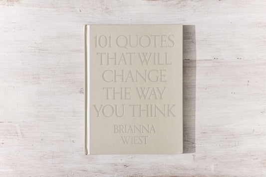 Beige hardcover book titled '101 Quotes That Will Change The Way You Think' by Brianna Wiest on rustic white wood table. Come see us at Awaken, Franklin, Tennessee