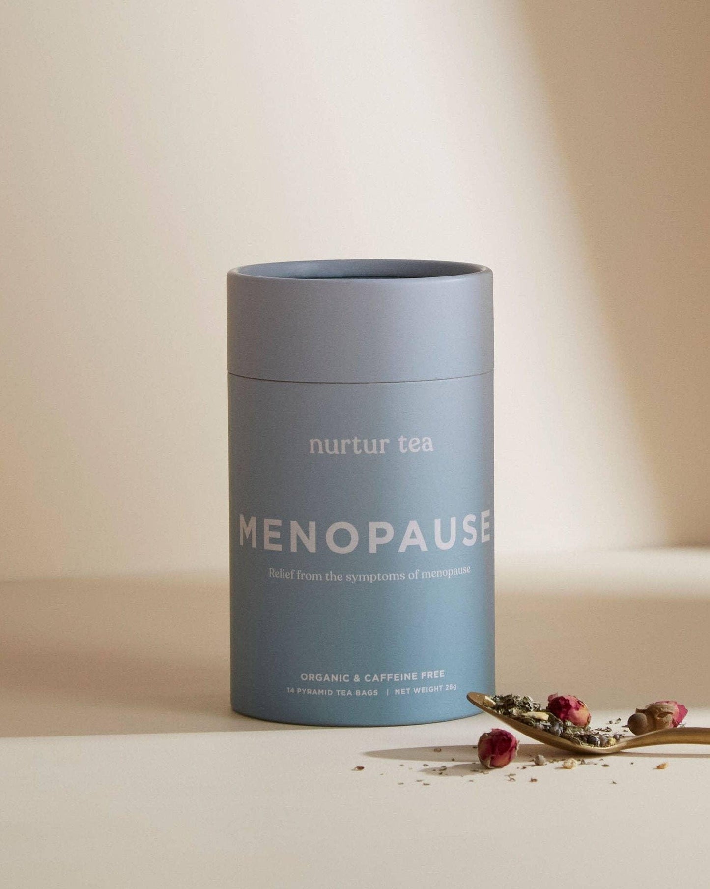 Blue Nurtur Tea Menopause relief container with dried herbs on spoon, minimalist beige background. Come see us at Awaken, Franklin, Tennessee