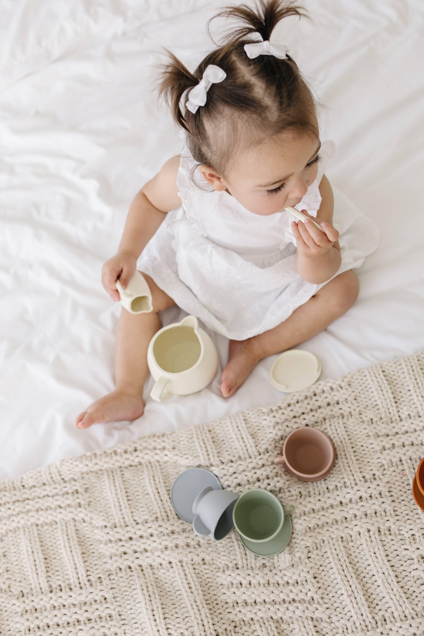 Toddler in white dress with pigtails playing tea set on bed, Come see us at Awaken, Franklin, Tennessee