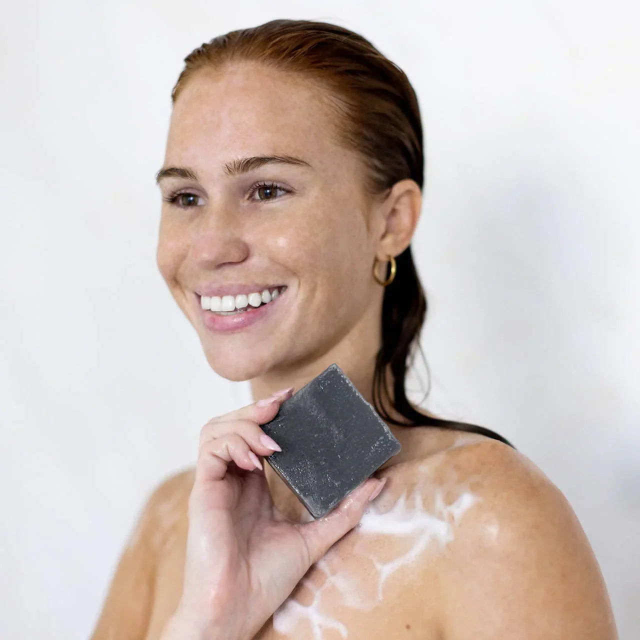Smiling woman with wet hair holding black soap bar with lather on shoulder, bright background. Come see us at Awaken, Franklin, Tennessee