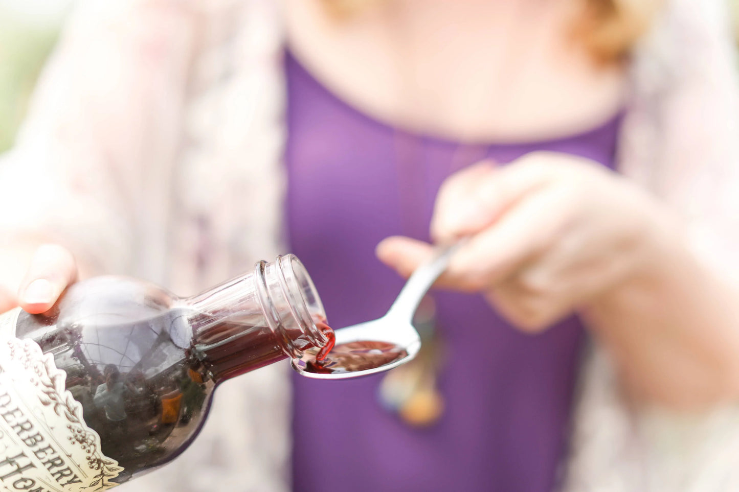 Person pouring dark elderberry syrup into spoon, wearing purple top and light cardigan, Come see us at Awaken, Franklin, Tennessee