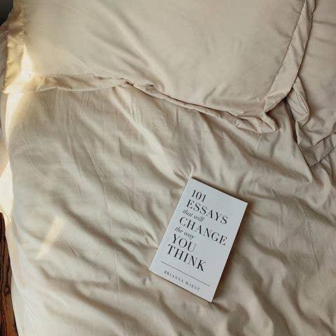 Book titled '101 Essays That Will Change The Way You Think' on beige bed linen with natural light. Come see us at Awaken, Franklin, Tennessee