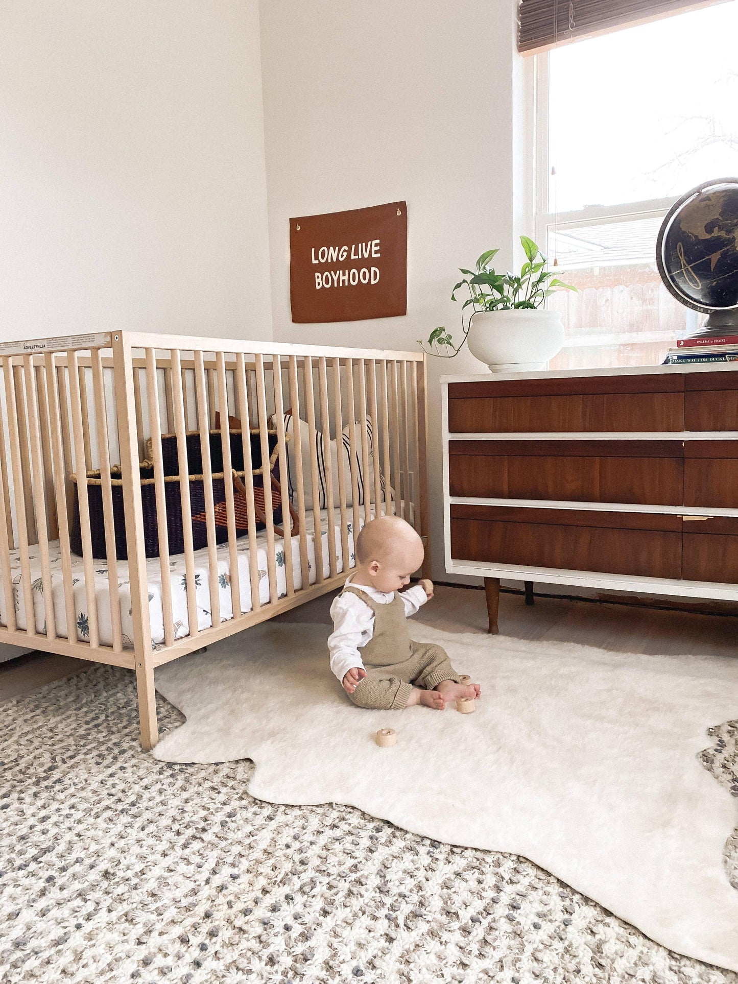 Minimalist nursery with baby sitting on white rug, wooden crib, dresser, and a sign saying LONG LIVE BOYHOOD. Come see us at Awaken, Franklin, Tennessee.