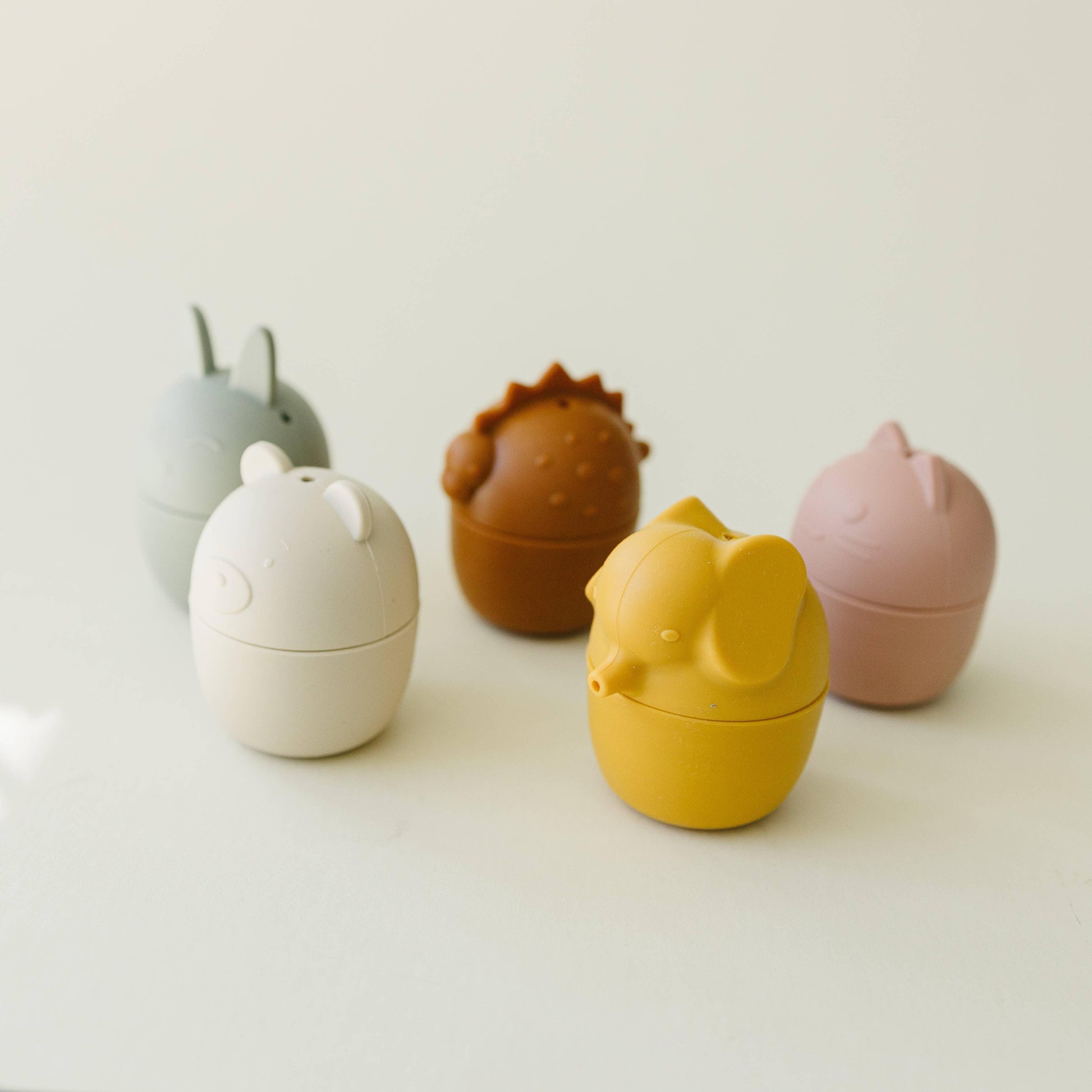 Yellow elephant, pink cat, brown dinosaur, white bear, and gray rabbit silicone containers on white surface. Come see us at Awaken, Franklin, Tennessee