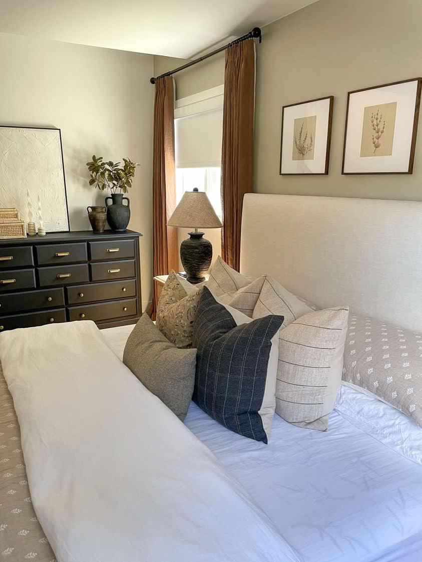 Cozy bedroom with decorative throw pillows, beige upholstered bed, black dresser, and lamp. Come see us at Awaken, Franklin, Tennessee