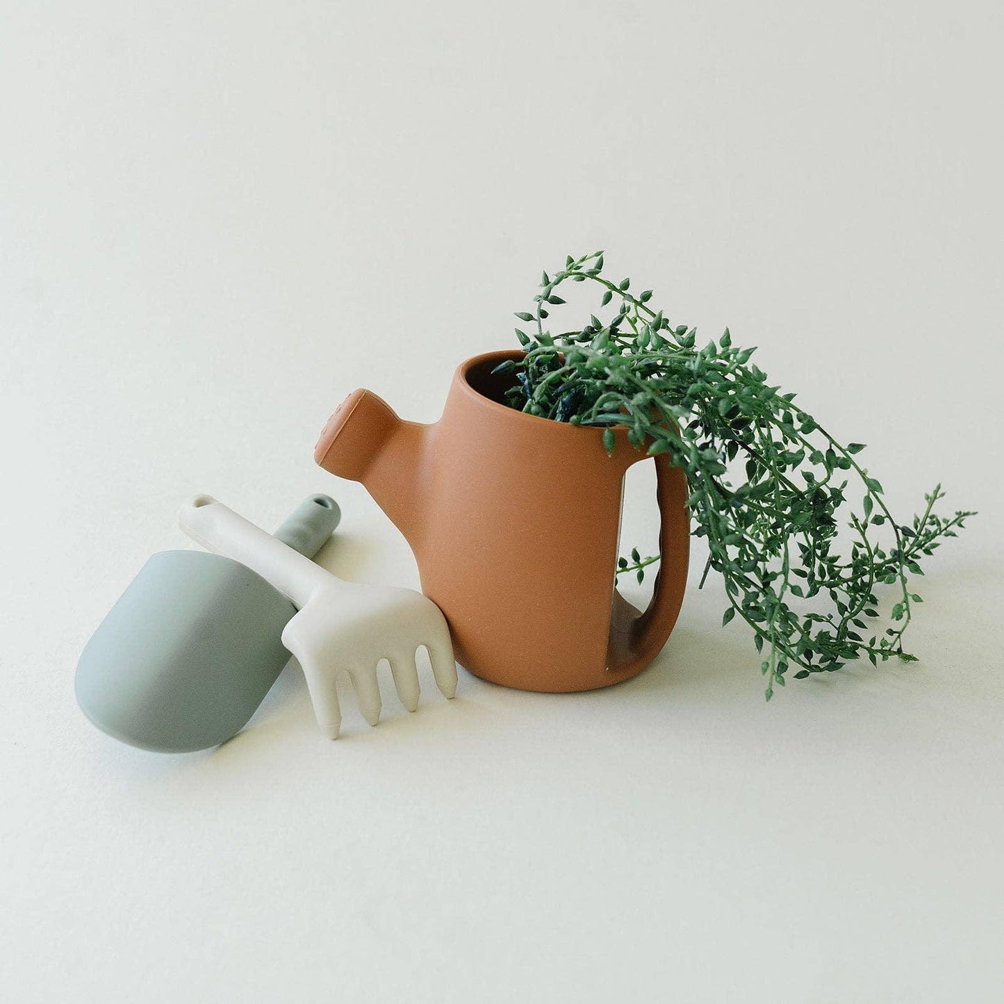 Terracotta watering can with green trailing plant and gray garden tools on white surface. Come see us at Awaken, Franklin, Tennessee