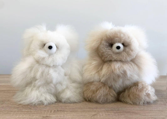 Two fluffy alpaca wool plush toys, one white and one beige, on wooden surface. Come see us at Awaken, Franklin, Tennessee
